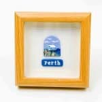 A small framed artwork featuring a depiction of a blue house surrounded by native plants under a bright sky with white clouds. Below the illustration, a blue plaque displays the word “Perth” in white lettering. The piece is set within a light wooden frame against a white background, creating a clean, modern presentation.