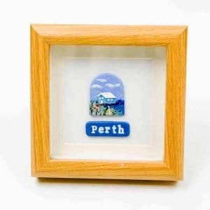 A small framed artwork featuring a depiction of a blue house surrounded by native plants under a bright sky with white clouds. Below the illustration, a blue plaque displays the word “Perth” in white lettering. The piece is set within a light wooden frame against a white background, creating a clean, modern presentation.