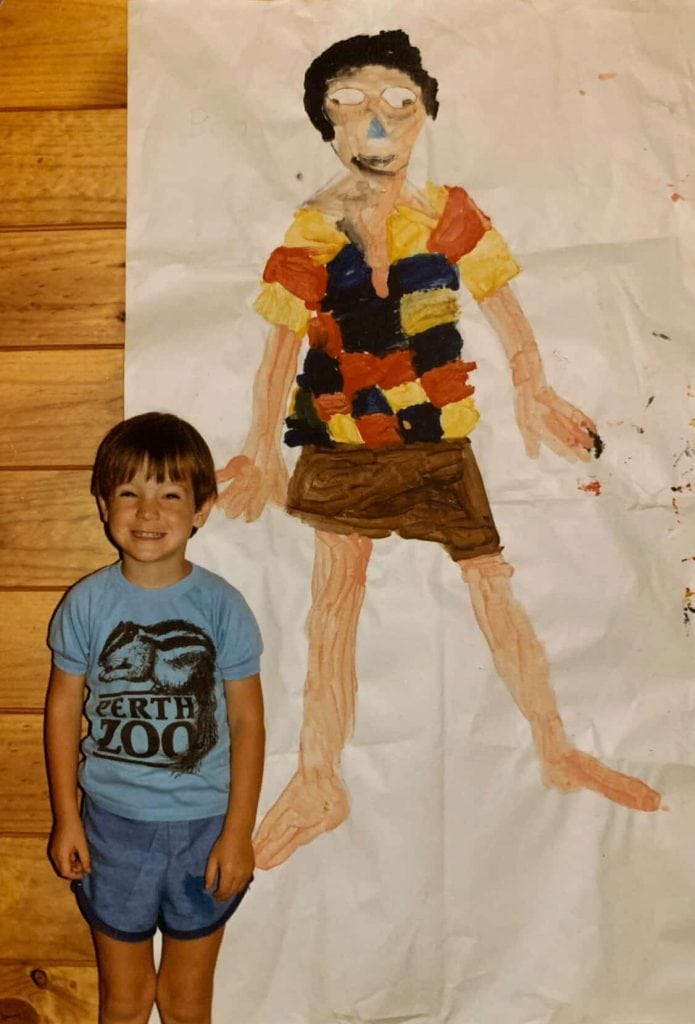 A young Ben Sherar as a smiling toddler wearing a blue Perth Zoo t-shirt, standing next to a large, colorful life-sized self-portrait painting traced and painted on butcher paper.