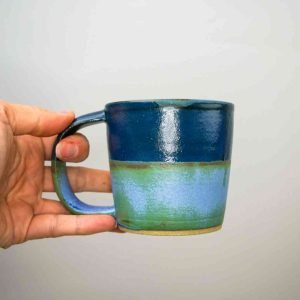 A fair-skinned hand holding a mug. The bottom of the mug is green and the top half is blue.
