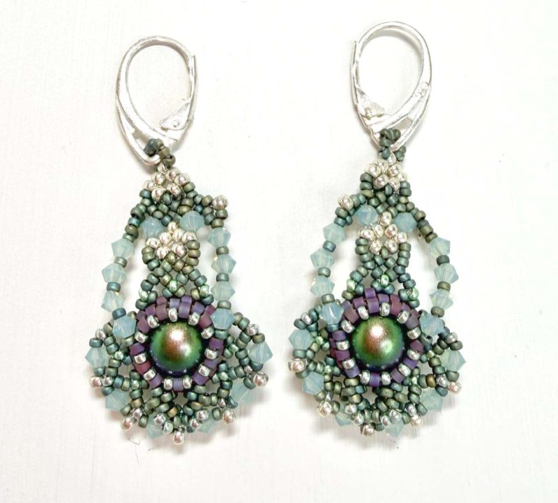 A pair of handcrafted beaded earrings featuring an intricate teardrop design. Each earring has a shimmering iridescent centre bead surrounded by small glass beads in shades of sage green, lilac, and pearl. The earrings are finished with silver lever-back hooks and arranged against a plain white background, highlighting their detailed texture and elegant craftsmanship.