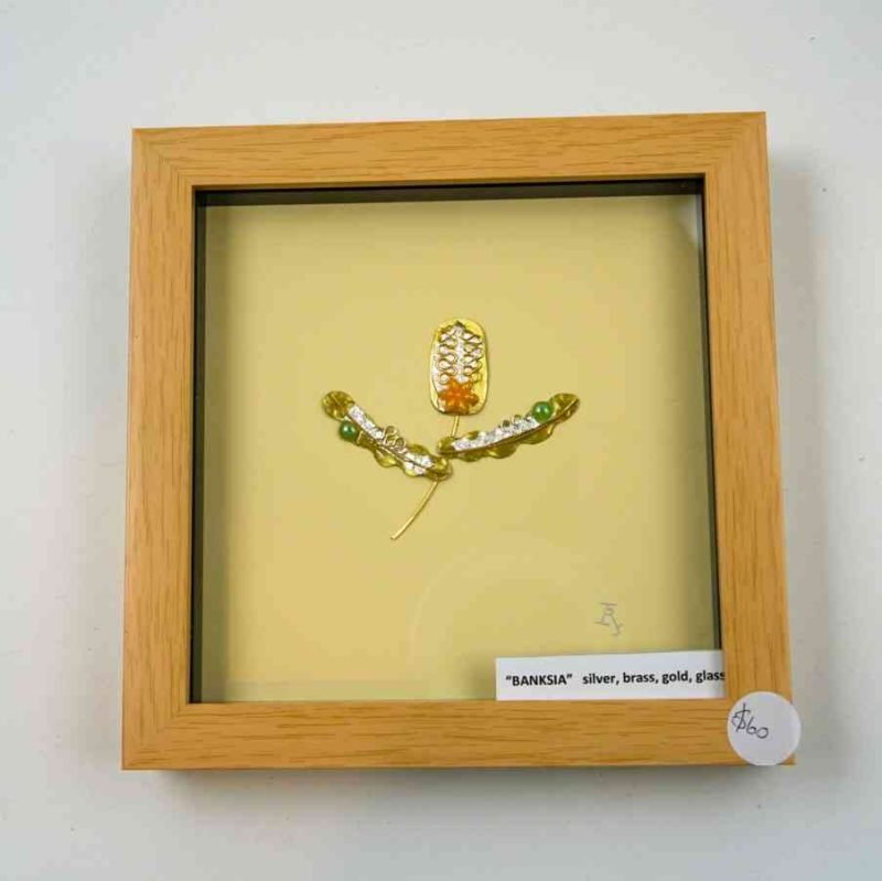 A wooden frame showcasing a silver scultpure of a banksia flower sits against a white background. The flower is made of silver, brass, gold, and glass beads