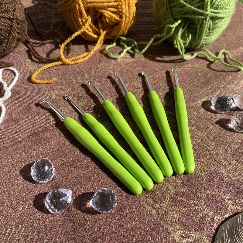 A group of 6 green crochet hooks are splayed across a brown patterned background, and surrounded by yarn, reflective crystals, and shells