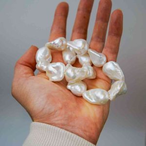 A hand holding several large, irregularly shaped white baroque pearls with a lustrous surface. The pearls vary in size and contour, reflecting light softly against a neutral grey background. The person’s sleeve is light beige, adding warmth to the composition.