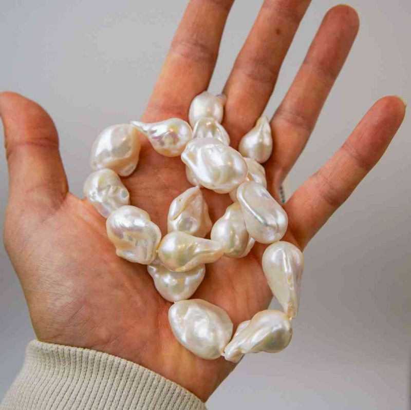 A hand holding several large, irregularly shaped white baroque pearls with a lustrous surface. The pearls vary in size and contour, reflecting light softly against a neutral grey background. The person’s sleeve is light beige, adding warmth to the composition.