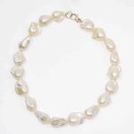 Jumbo Pearl Strand Colleen Brennan (3) A close-up image of a necklace made from irregularly shaped white baroque pearls arranged in a circle. Each pearl has a lustrous, organic surface with subtle iridescent tones, and the strand is finished with a small silver lobster clasp. The background is plain and light, highlighting the pearls’ natural sheen and texture.