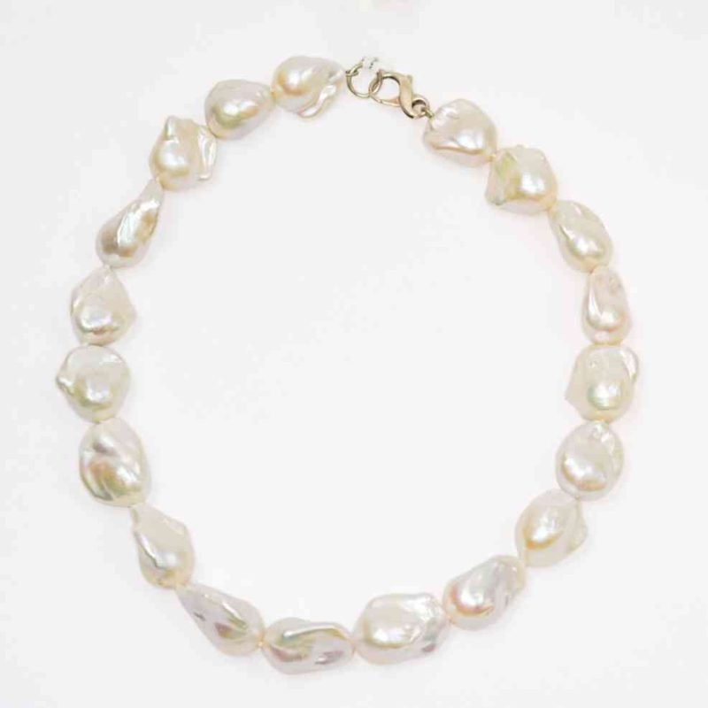 A close-up image of a necklace made from irregularly shaped white baroque pearls arranged in a circle. Each pearl has a lustrous, organic surface with subtle iridescent tones, and the strand is finished with a small silver lobster clasp. The background is plain and light, highlighting the pearls’ natural sheen and texture.