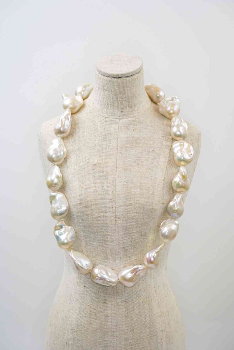 A beige linen mannequin displaying a necklace made of large, irregularly shaped white baroque pearls. Each pearl has a lustrous, organic surface with soft iridescent tones of cream and silver. The necklace drapes naturally around the mannequin’s neck, highlighting the unique size and texture of each pearl against the neutral background.