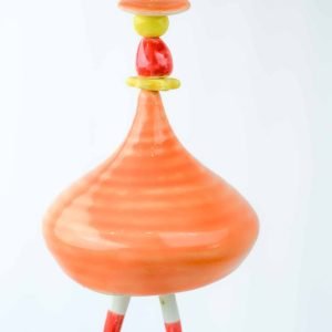 An orange wind chime in the shape of a ballerina hangs before a white background. The bell is a tapred shape with two stick-shaped 'legs' as the tongue of the bell. The head of the ballerina is made of a red gumnut-shaped bead.