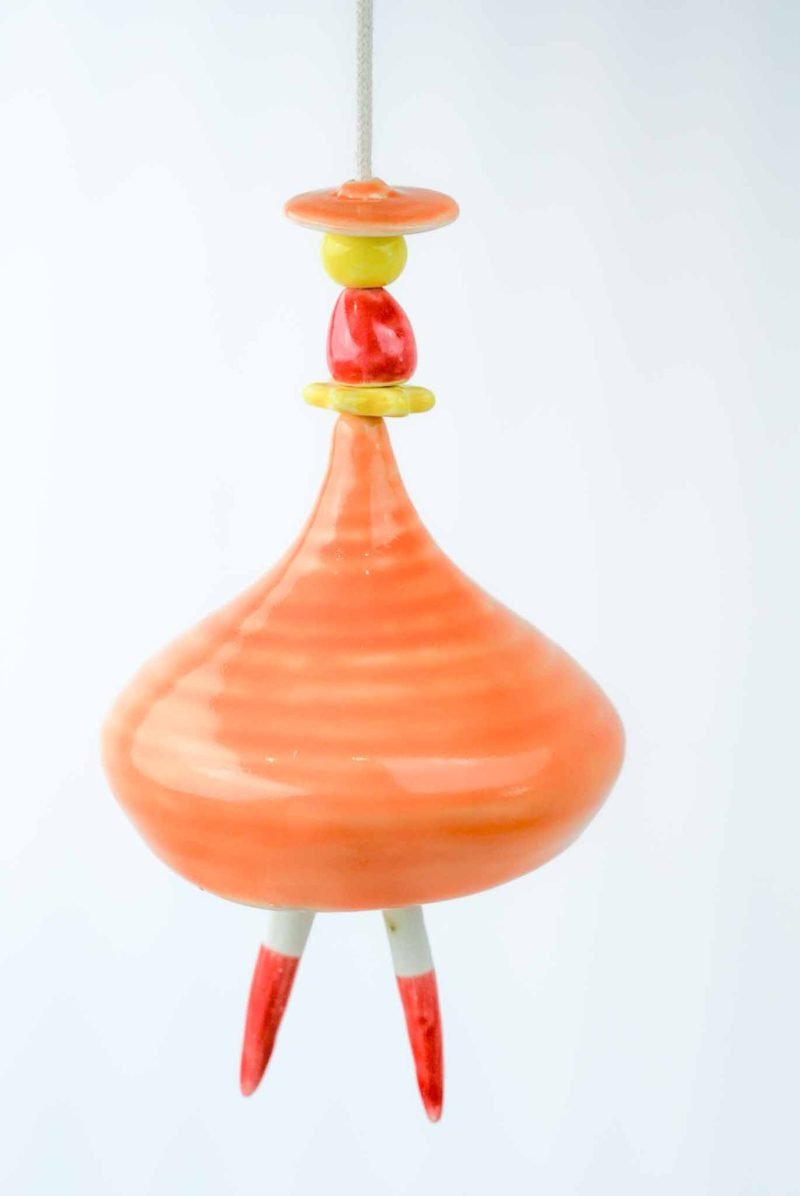 Large Ballerina Porcelain Bell Belen (3) An orange wind chime in the shape of a ballerina hangs before a white background. The bell is a tapred shape with two stick-shaped 'legs' as the tongue of the bell. The head of the ballerina is made of a red gumnut-shaped bead.