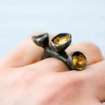 An oxidised silevr ring is worn on the hand of a fair-skinned woman. The ring has 3 seed-pod accents atop it, lined with gold to resemble to seed pods in nature the design is taken for inspiration