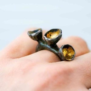 An oxidised silevr ring is worn on the hand of a fair-skinned woman. The ring has 3 seed-pod accents atop it, lined with gold to resemble to seed pods in nature the design is taken for inspiration