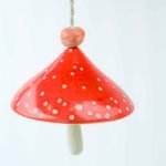 A ceramic wind chime hangs against a white background. The wind chime is of a toadstool, with the bell of the wind chime glazed bright red with white spots, and the tongue of the bell made to be the foot of the toadstool.