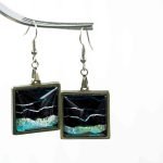 Square Dangler Earrings Noetic Domain (1) A pair of square earrings made from resin set in silver frames hangs from a fork on a white background. The square earrings are filled with black and sparkly blue abstract patterns