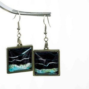 A pair of square earrings made from resin set in silver frames hangs from a fork on a white background. The square earrings are filled with black and sparkly blue abstract patterns