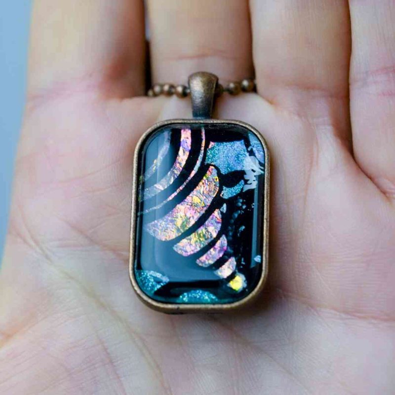 The rectangular pendant lays on the open palm of a fair-skinned woman, showcasing the size. The pendant rests from the base of her finger down to the second line in her palm.