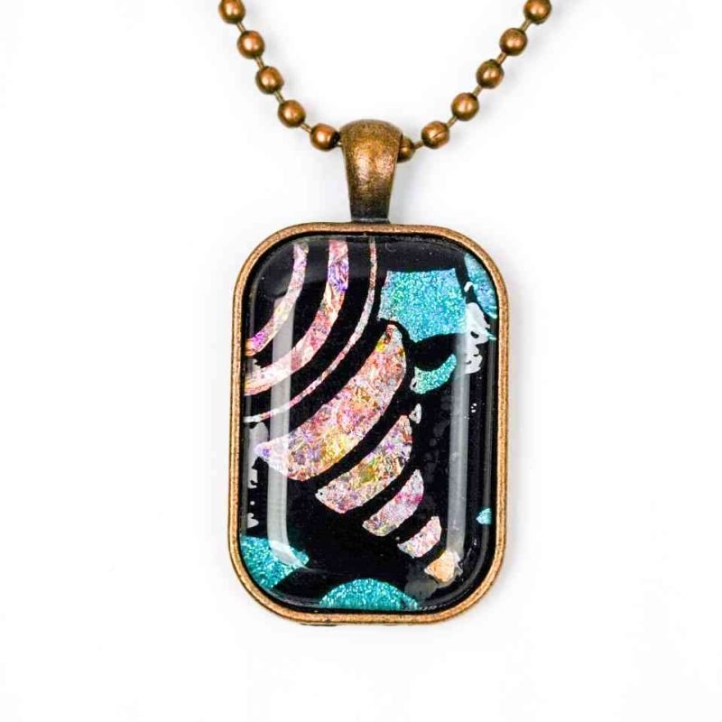 rectangle pendant (2) A Large rectangular pendant hangs on a copper-toned chain, resting against a white background. The pendant is made up of a copper-toned frame filled with resin. The resin is clear and covers an abstract design consisting of a black base, and an opalescent, peach-coloured conical shape resembling a spire shell