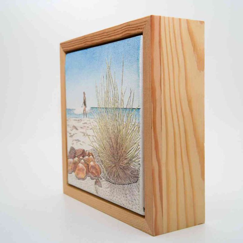 Framed ballpoint pen drawing of a beach scene showing rocks and grass in the foreground, with a person walking along the shoreline under a clear blue sky.