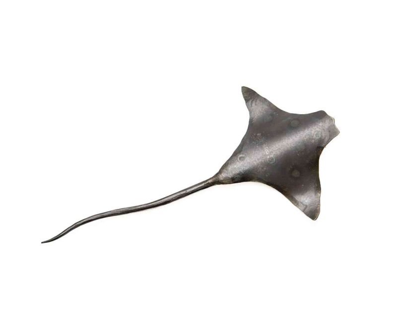 A metal sculpture of an eagle ray lays on an all white background. The ray is made of a dark metal and has some subtle circles on the back to resemble an eagle ray