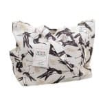 Magpie Song Tote bag (1) A cream-coloured canvas tote bag featuring a bold, stylised print of Magpies with sharp beaks and red eyes, accented by soft golden botanical motifs. The bag has sturdy handles made from the same patterned fabric, roomy interior space, and side pockets for extra storage.