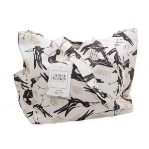 A cream-coloured canvas tote bag featuring a bold, stylised print of Magpies with sharp beaks and red eyes, accented by soft golden botanical motifs. The bag has sturdy handles made from the same patterned fabric, roomy interior space, and side pockets for extra storage.