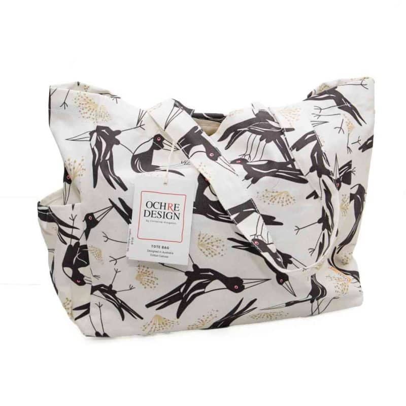Magpie Song Tote bag (1) A cream-coloured canvas tote bag featuring a bold, stylised print of Magpies with sharp beaks and red eyes, accented by soft golden botanical motifs. The bag has sturdy handles made from the same patterned fabric, roomy interior space, and side pockets for extra storage.