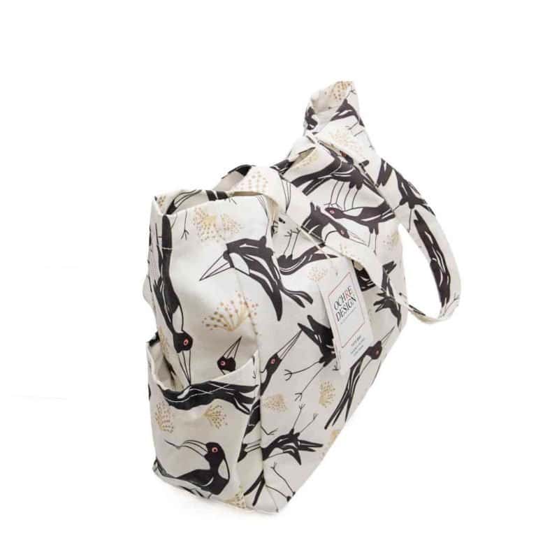 A cream-coloured canvas tote bag featuring a bold, stylised print of Magpies with sharp beaks and red eyes, accented by soft golden botanical motifs. The bag has sturdy handles made from the same patterned fabric, roomy interior space, and side pockets for extra storage.