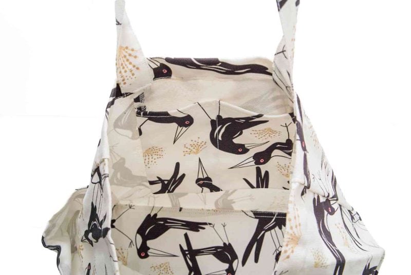 A cream-coloured canvas tote bag featuring a bold, stylised print of Magpies with sharp beaks and red eyes, accented by soft golden botanical motifs. The bag has sturdy handles made from the same patterned fabric, roomy interior space, and side pockets for extra storage.